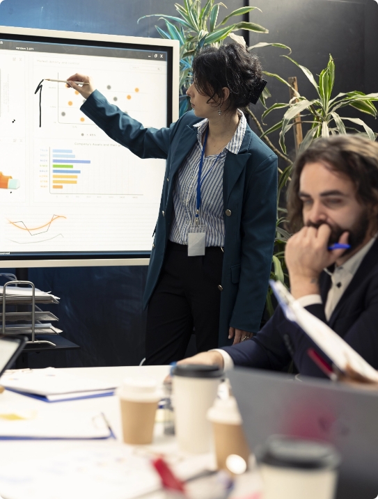 A professional woman giving a presentation using a large digital display showing various business charts and analytics to a colleague in a modern office.